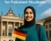 A smiling Pakistani student holding a German flag in front of a historic German university building, promoting DAAD 2025 scholarships.